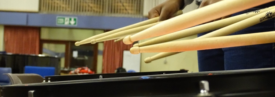The bottom of the photo is a close up of the rim of a marching snare drum. The rim of a second drum is visible behind the first. The hands and of someone wearing a grey hoodie and blue jeans are holding a set of drumsticks over the back drum. in fron of this person there are two more pairs of sticks being held over the line of drums though the holders are not visible.